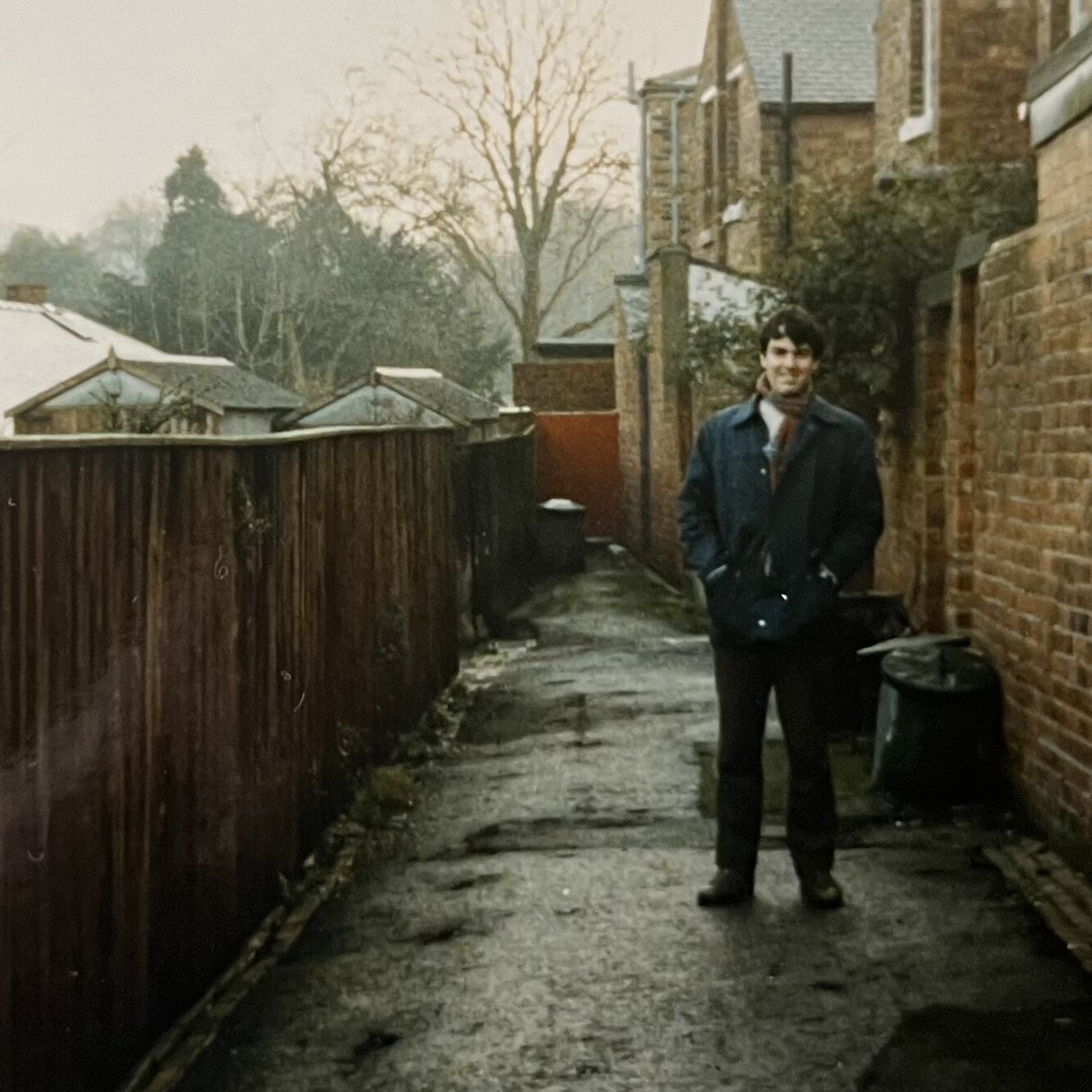 A man standing at the end of a wet back lane between brick houses and wooden fences